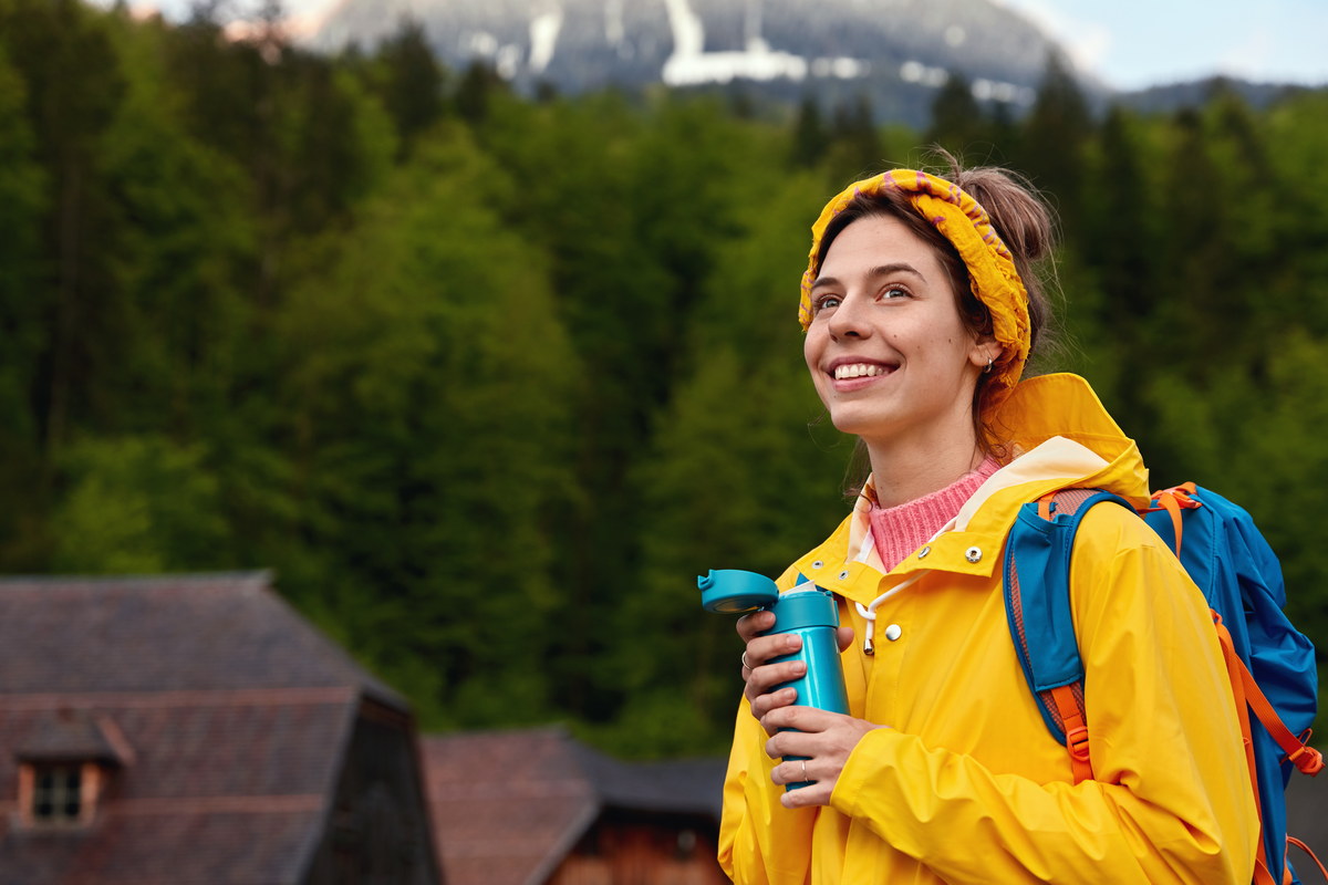 cheerful lovely woman with pleasant smile, drinks tea from thermos, spends holidays in mountain resort, poses in calm place against green coniferous forest small houses. people, rest, nature concept