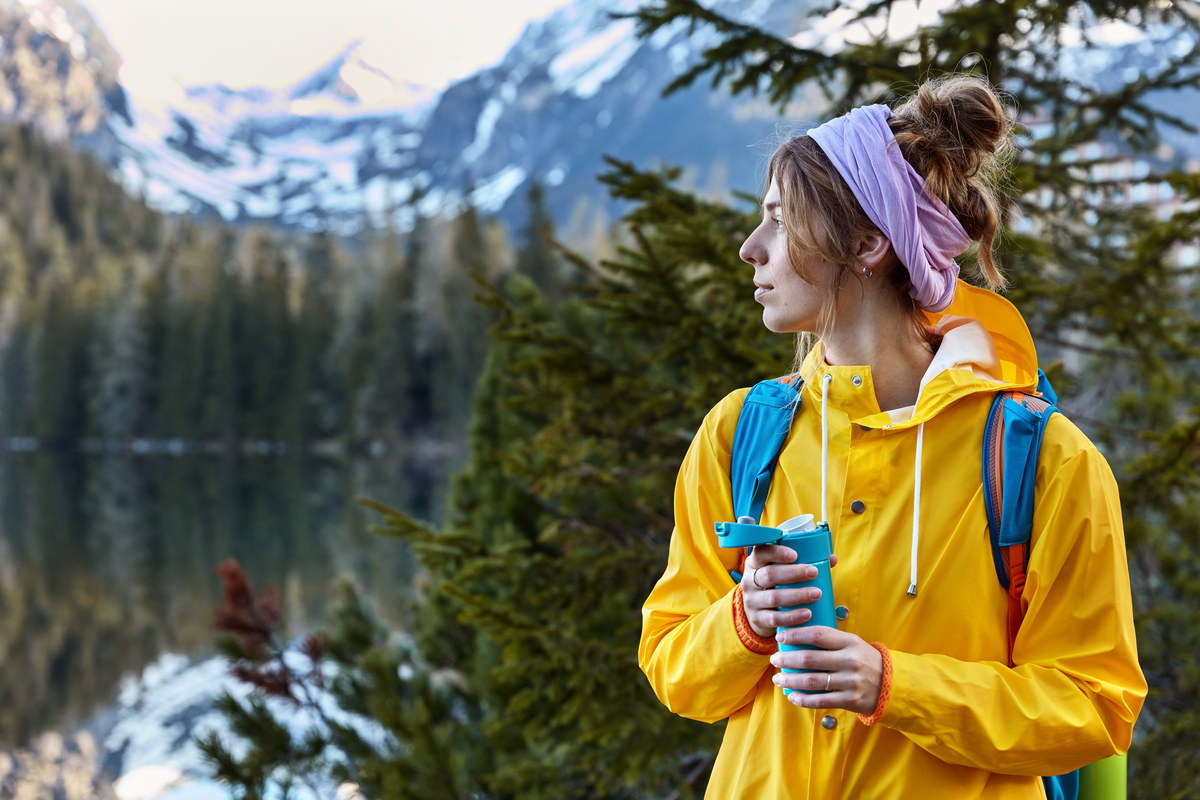 pensive female mountaineer wears scarf around head, yellow raincoat, enjoys hot coffee from flask, has trip in mountains, poses on nature landscape with copy space, hikes in beautiful forest