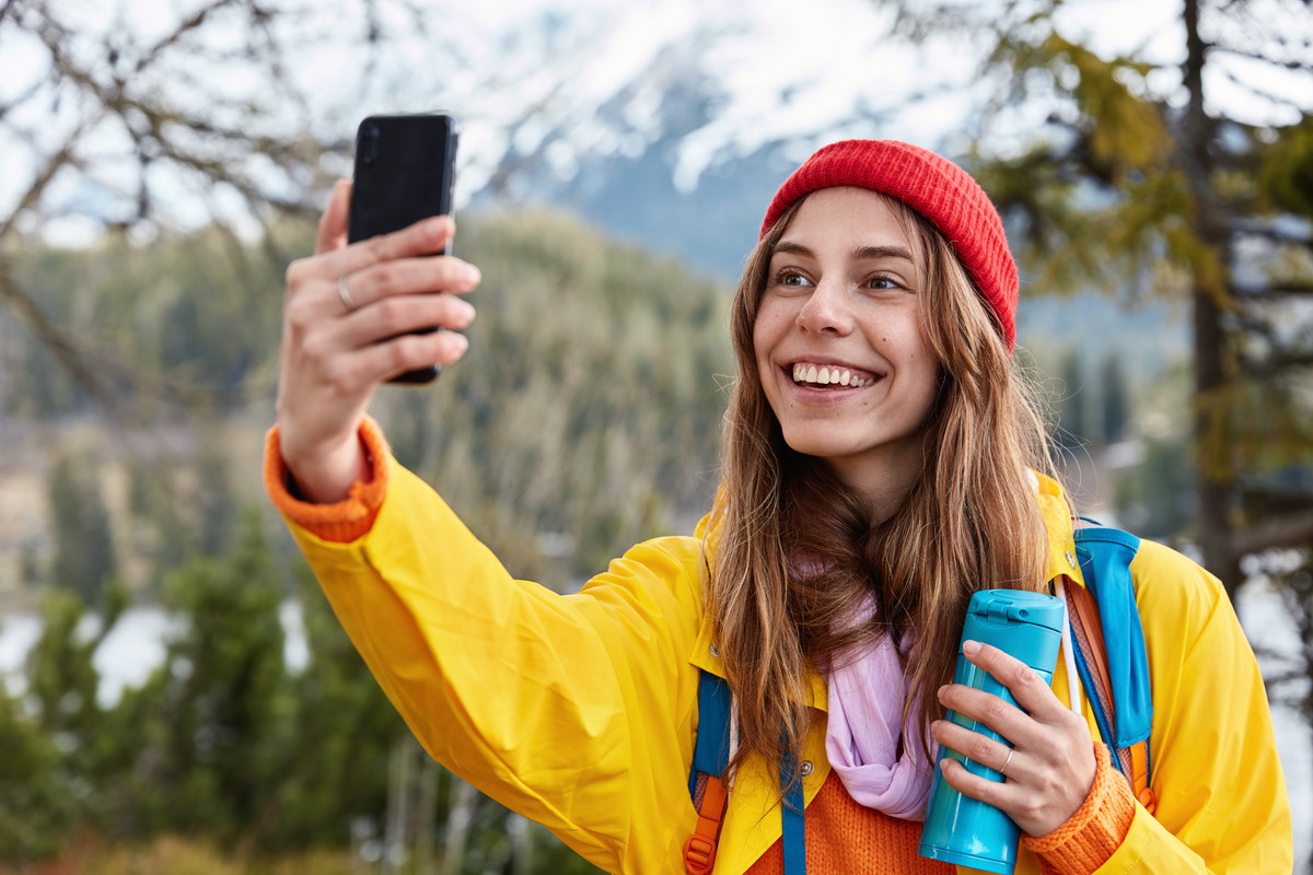 smiling female tourist holds smart phone, takes selfie against beautiful landscape of coniferous wood, holds thermos of hot drink, wears red hat and yellow coat, walks through forest. people, journey